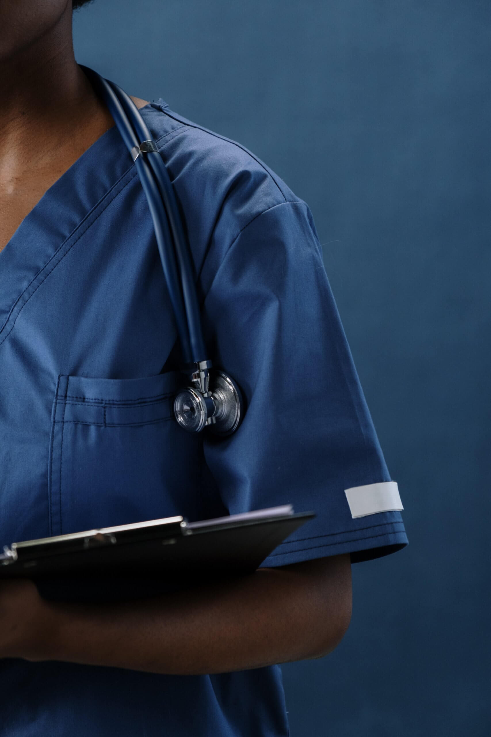 Dottoressa di carnagione scura con camice blu, stetoscopio al collo e una cartelletta con un foglio.