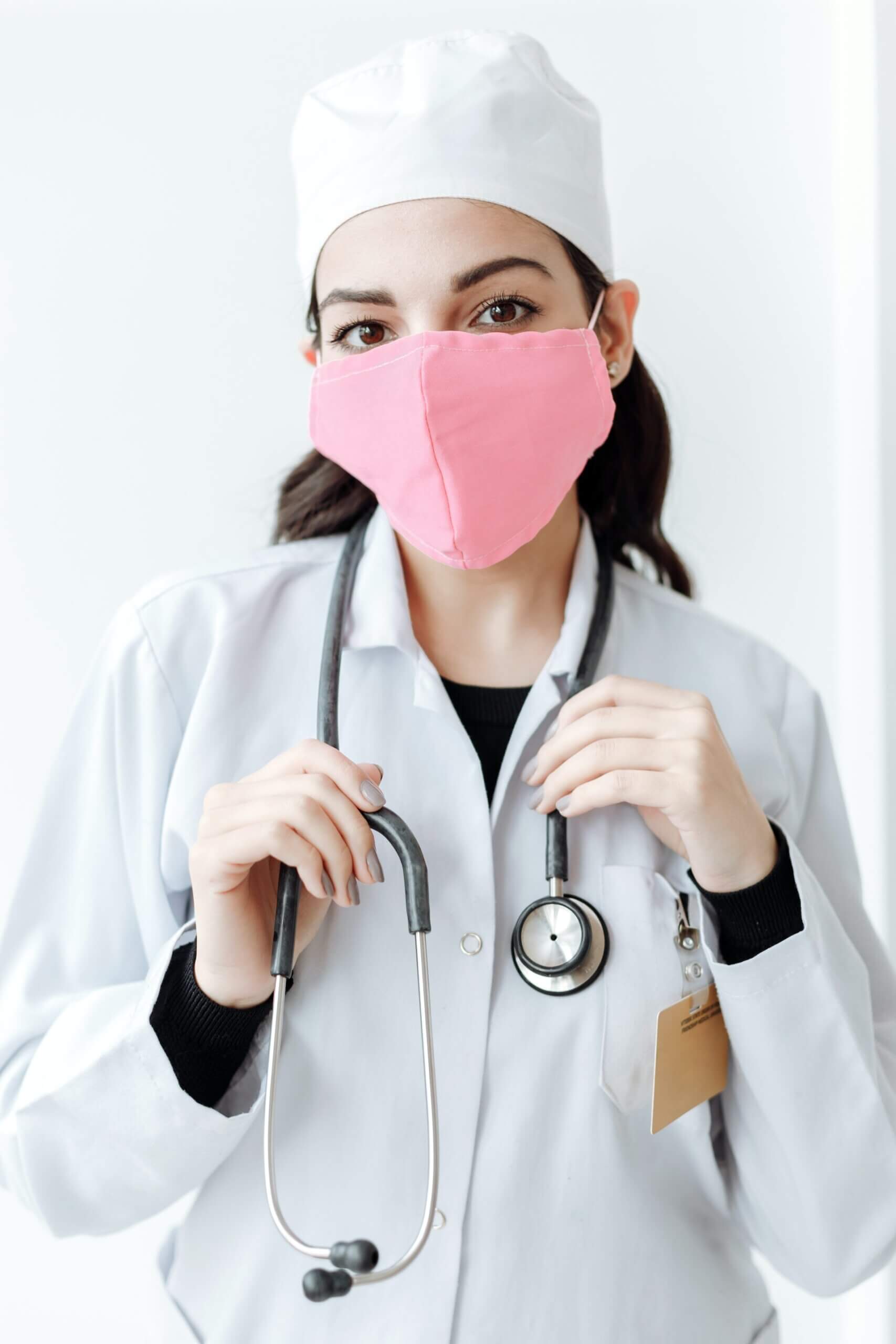 Infermiera donna con copricapo bianco, mascherina rosa, camice bianco, stetoscopio al collo e tesserino sulla tasca alta del camice che guarda dritto nell'obbiettivo della fotocamera.